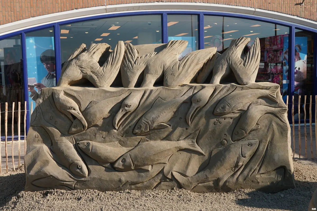 Escultura de arena con aves en la parte superior y peces tallados debajo.