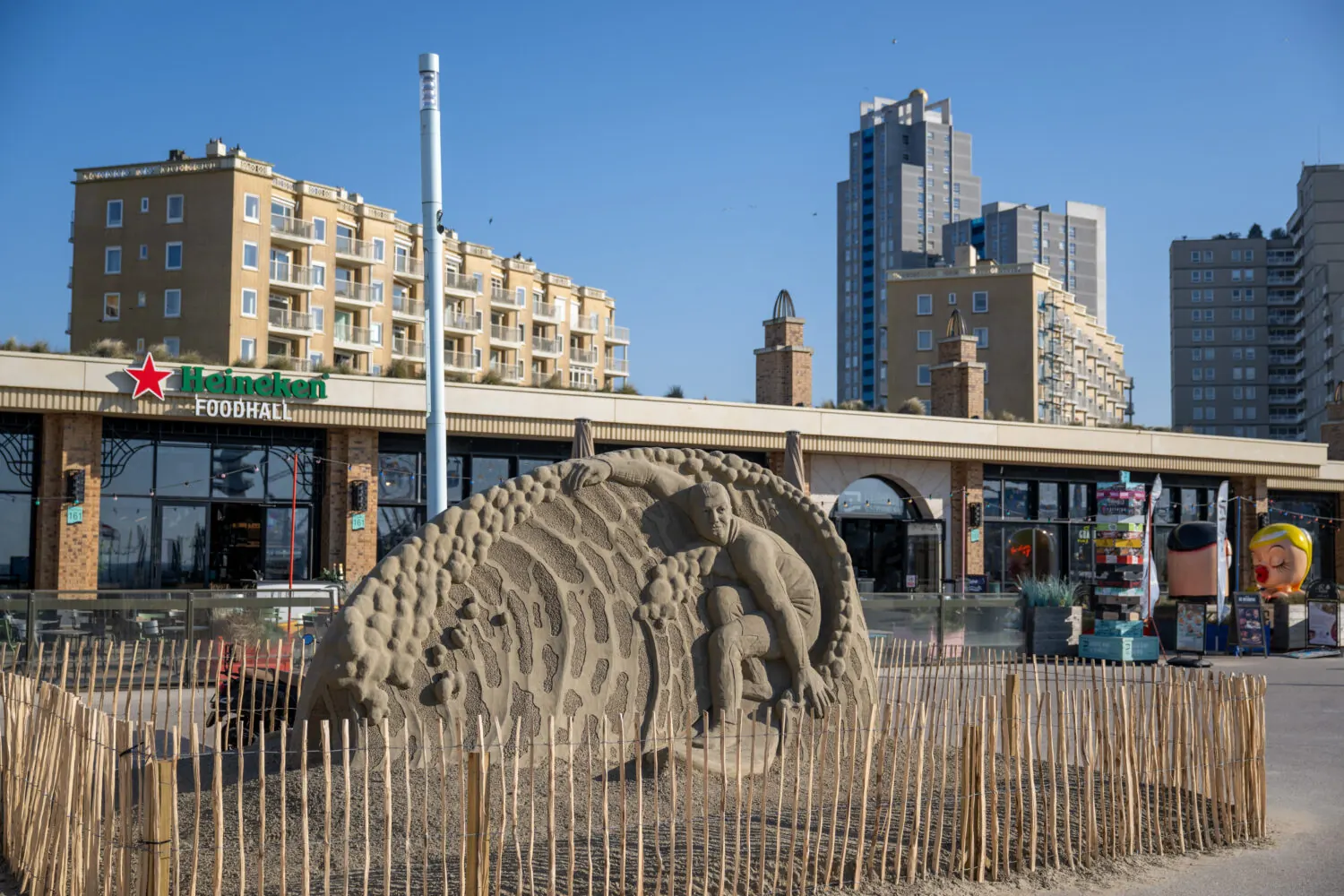 Escultura de arena con una figura sobre una ola, situada frente a un paseo marítimo.